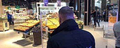 Sicherheitsmitarbeiter mit Jacke „proSicherheit“ beobachtet Verkaufsfläche mit Obst- und Gemüseständen im Markt