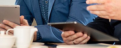 Zwei Personen am Tisch besprechen Unterlagen, eine Person hält ein Tablet, daneben Laptop und Notizbuch