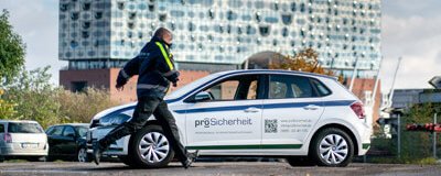 Mitarbeiter geht neben einem ProSicherheit-Fahrzeug, im Hintergrund ein Gebäude mit Glasfassade.