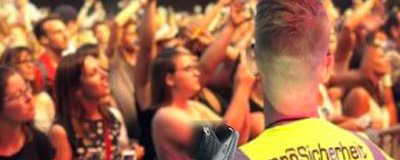 Sicherheitsmitarbeiter in Warnweste mit Funkgerät steht vor einer Menschenmenge bei einer Veranstaltung