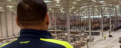 Sicherheitsmitarbeiter in Jacke mit Logo steht in einer Lagerhalle und blickt auf Regale und Arbeitsbereich