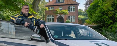 Sicherheitsmitarbeiter lehnt an einem Dienstfahrzeug vor einem Wohnhaus, umgeben von Bäumen.