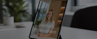 Tablet auf einem Schreibtisch zeigt Willkommensbildschirm mit Frau und Text „Herzlich Willkommen“