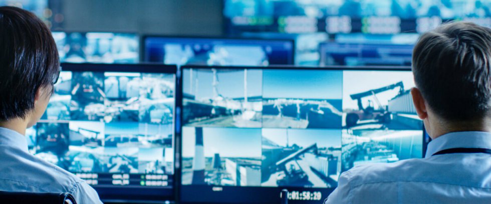 Zwei Mitarbeitende von hinten vor Monitoren mit mehreren Videoüberwachungsbildern in einer Leitstelle