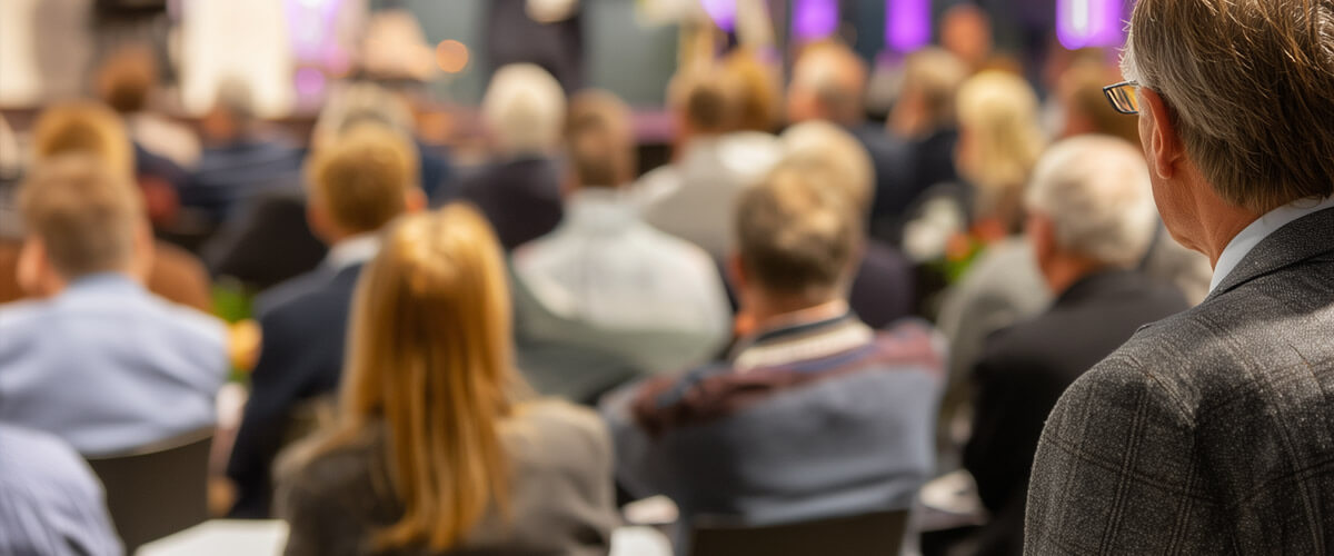 Publikum in einem Veranstaltungssaal, rechts eine Person im Anzug von hinten, im Hintergrund Bühne unscharf